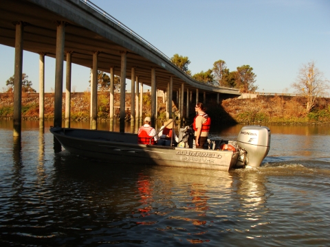 Three people in a motorized boat pass under a bridge in a river
