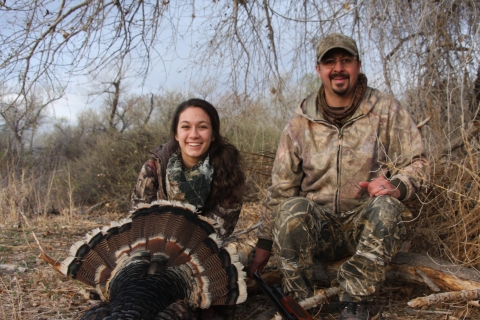 A young woman and a man in camouflage sit on a ground in a wooded area with a wild turkey