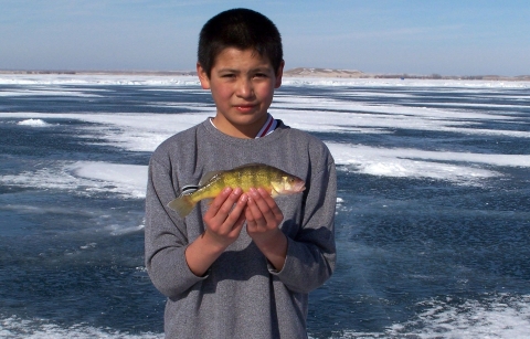 A young boy standing on a frozen, snow-swept lake holding a small fish in both of his hands
