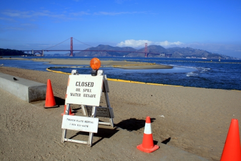 A sign on a small barrier says "Closed, Oil Spill, Water Unsafe" with San Francisco Bay in the background.