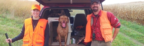 A woman, her husband and their dog standing near the tailgate of their car in hunting gear