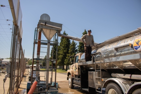 A man stands atop a truck with a pipe feeding into it from a tall metal structure