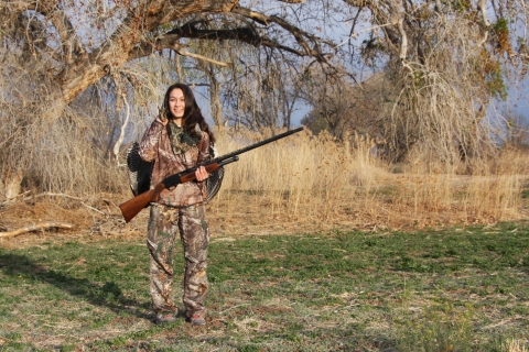 A young woman in camouflage holds a turkey over her shoulder and a gun across the front of her body. She is standing in a wooded area.