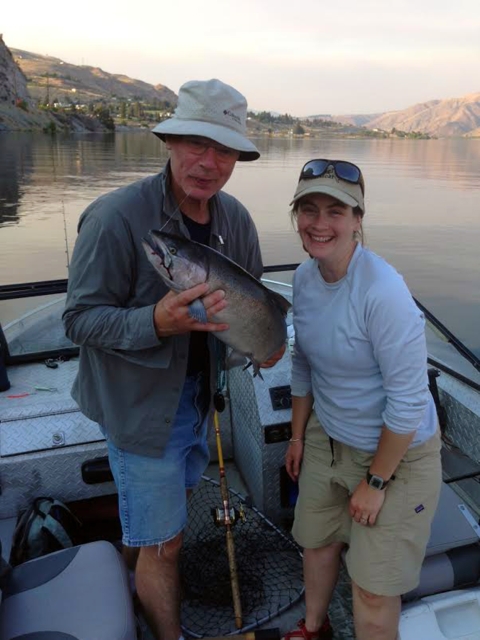 A man and woman pose grinning in the back of a boat, the man holding up a big salmon. Both people wear shorts and sun hats.