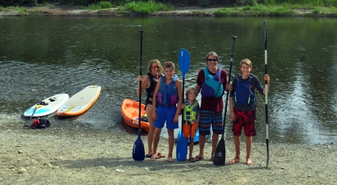 A family with mother, father, and 3 sons pose in front of their paddleboards in the river, holding their paddles and wearing lifejackets.