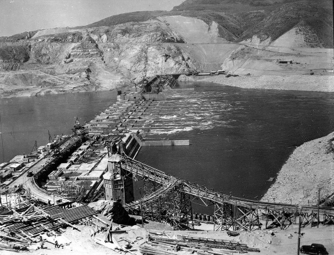 Black and white photo of Grand Coulee Dam under construction, showing infrastructure spanning the river but not yet completely blocking flow.