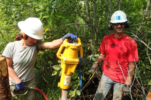 A woman and man in t-shirts, hardhats, and ear protection muffs stand in a thicket of brush, both smiling. The woman holds a mechanical device upright with one hand and a hydraulic hose in the other and is looking down at what the device is doing out of the photo's frame.
