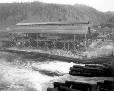 Black and white photo of an old sawmill perched on stilts beside a rushing river, with riverbank supported by logs and logs used to form a kind of dam in the foreground.