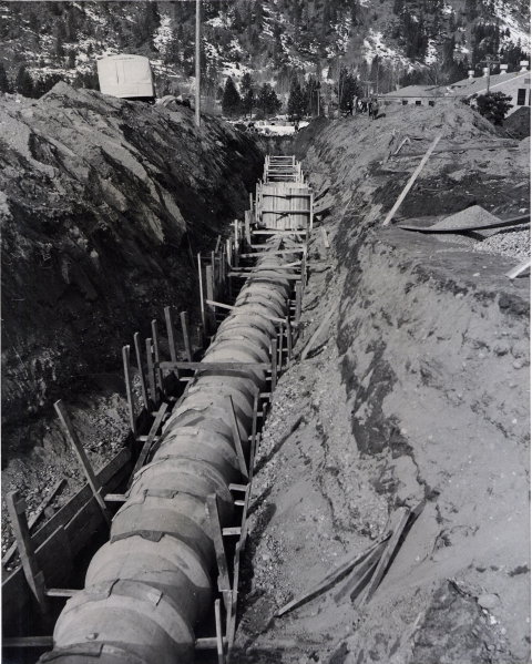 Black and white photo of a deep trench in sand and gravel, and a concrete pipe being laid, with a snowy mountainside visible in the background and a glimpse of hatchery buildings.