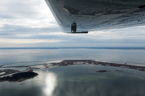 An airplane wing seen from inside the plane occupies the top center-right portion of the image. Below an island, visually similar to a long barrier island, is visible in the waters of Chesapeake Bay. 