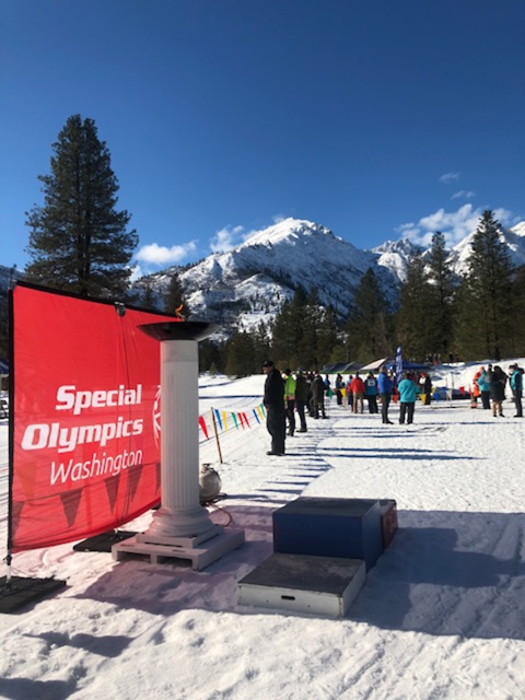 A bright sunny winter day. People are dressed warmly, watching a race from one side of a flagged barrier. In the foreground, a large banner reads, "Special Olympics, Washington" next to an eternal flame pedestal and three low platforms where the winners stand to receive medals.
