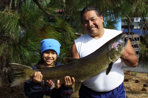 A boy in a blue hat and jacket and a mal in a white t-shirt smile as they hold up a huge salmon together, with pine boughs behind them.