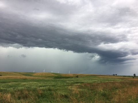 Rolling green and brown hills covered in prairie grasses receive much needed rain as a horseshoe shaped storm cloud passes overhead. A couple white wind turbines and green trees dot the horizon.