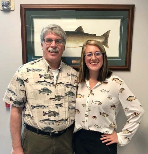 A man and a woman wearing shirts with fish print, standing in front of a framed photo of a fish