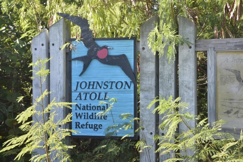 A wooden sign that reads Johnston Atoll National Wildlife Refuge. On the sign is a picture of e great frigatebird.