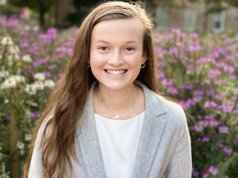 Young woman with long brown hair smiles with purple and white flowers in the background