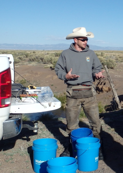A man wearing waders talking and standing next to an open truck bed with blue buckets at his feet
