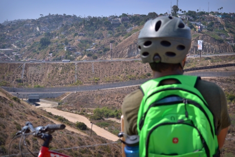 Youth mountain biker, back turned to the camera, wearing helmet and neon green backpack. A bikes' handlebars are visible on the bottom left of the photo. Youth is looking at mountain biking trails and urbanized area off in the distance. 