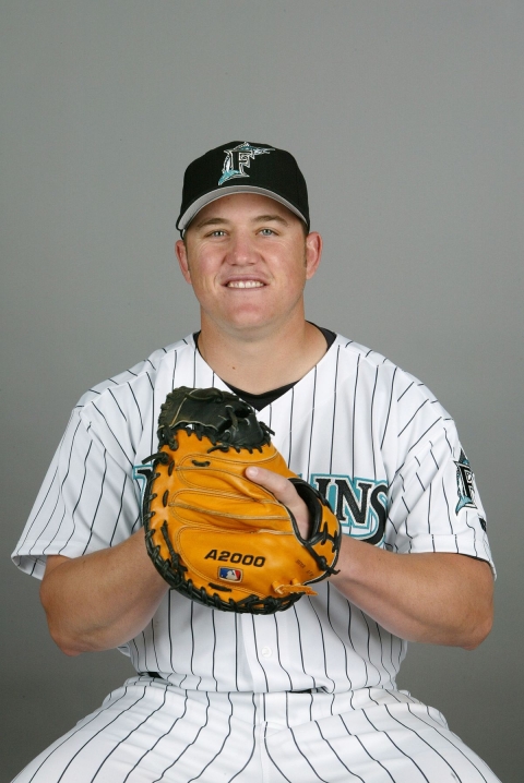 A young man in a baseball uniform and hat holds a leather baseball glove. He is against a grey background.