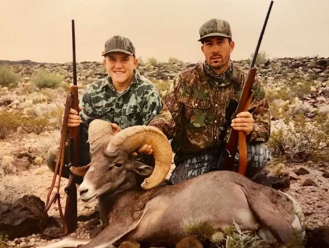 Two men wearing camouflage each hold a long gun and a bighorn sheep. They're in a rocky landscape with little vegetation. 