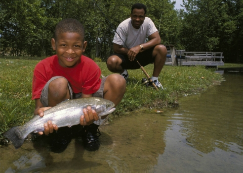 Young boy squatting on waters edge holding fish, man in background with fishing rod 