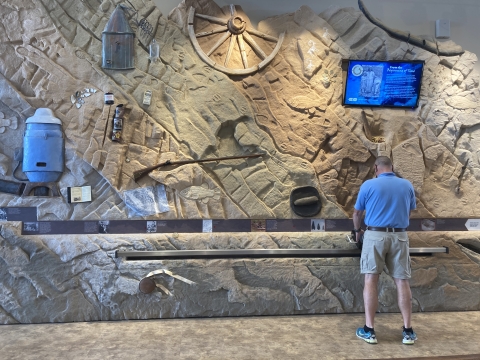 One man is standing infront of a timeline of artifacts on a wall with a television screen displaying information