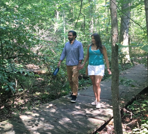 Bearded man and women walking on boardwalk through forest holding hands.