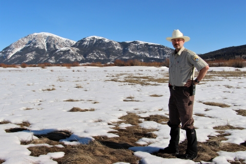 A man in a cowboy hat stands on a snowy landscape with mountains in the background.