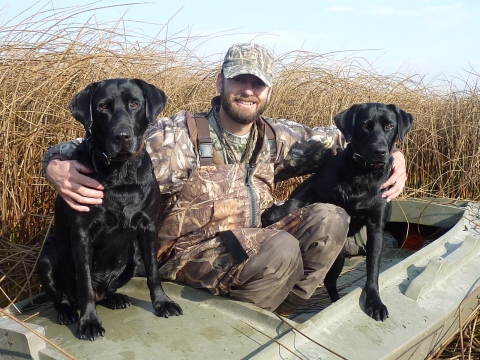A hunter and his two black labrador retrievers pose for a picture in the bulrushes of a lake
