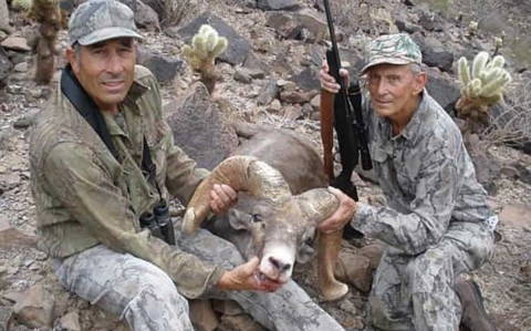 Two men wearing camouflage hold a bighorn sheep by its horns while sitting on the rocky ground