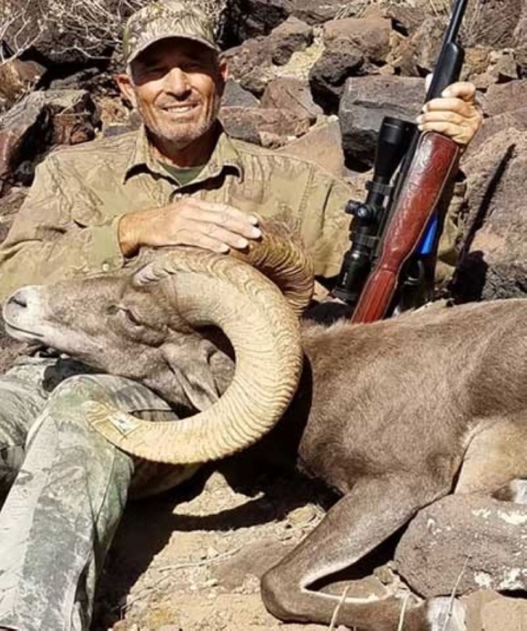A man in camouflage sits on a rocky ground with a bighorn sheep with large curling horns 