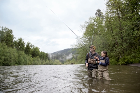 A parent and child stand in a river, fly fishing.