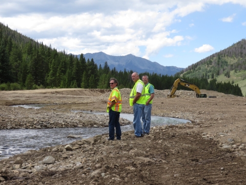 Three men in high visibility vests stand by river with forest in the background