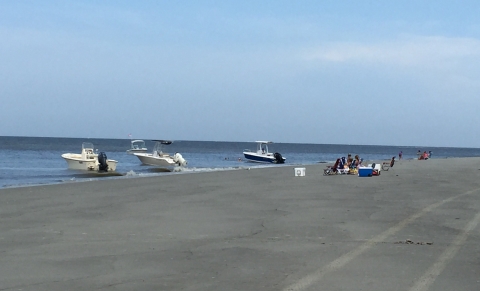 Three boats sit at the shoreline while people sit on the beach nearby. 