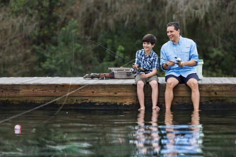 A boy and an adult sit on a dock by the water fishing. 
