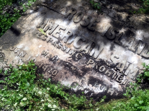 A worn plaque lays on the ground. Its word barely legible as the brush grows over it. Its only legible words read Rose Island American Samoa Trespassing Prohibited Jan 10, 1920