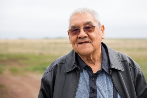 Elder Unangax man wearing grey jacket and blue shirt stands with blurred road and tundra in the background.