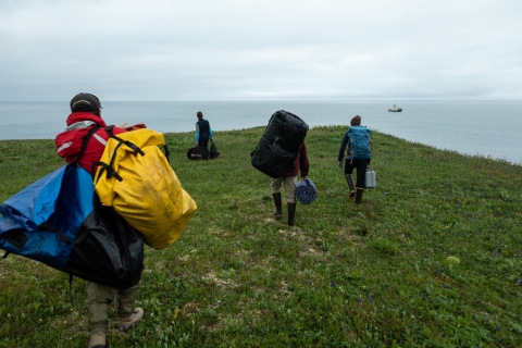 People carry large waterproof packs towards a green point of land overlooking the ocean. A blue and white ship is in the background.