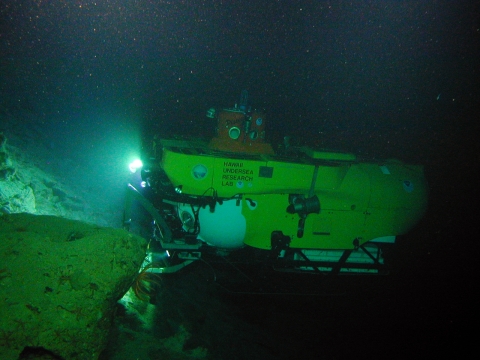 The Pisces V sub floats around some rocks. It is yellow and has a light shining. 