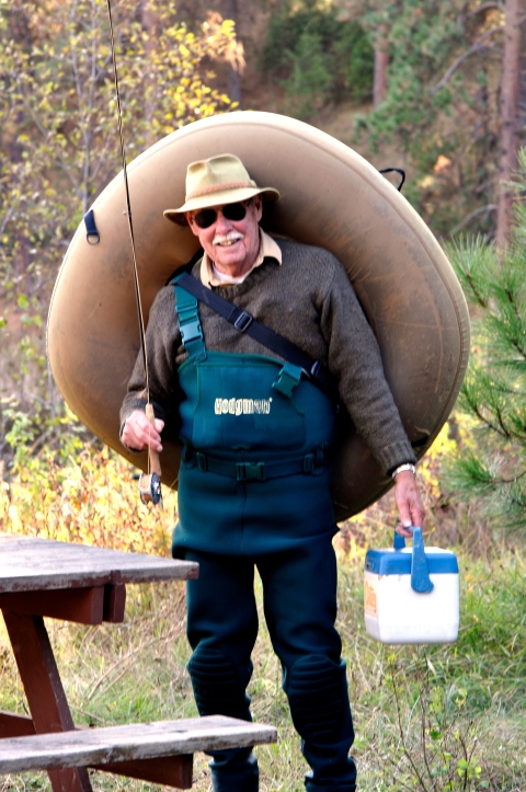 Man carrying a fishing inner-tube, rod and tackle box, wearing fishing fear and a big grin on his face.
