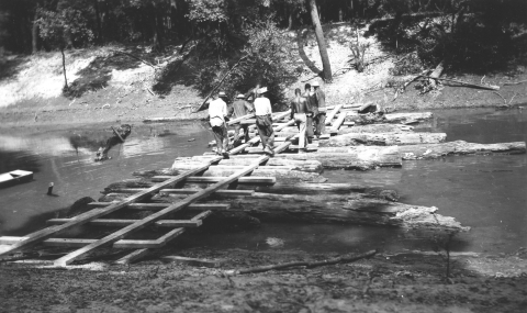 Black and white photo of five black men building a wooden bridge over floating logs