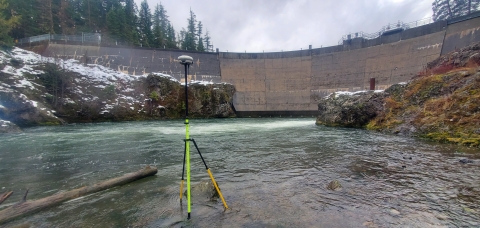 View of a dam from downstream with survey equipment set up in the river. Some snow on the riverbanks.
