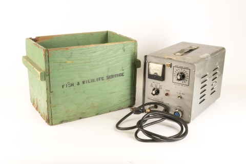 Upon a solid white background sits a handmade light green wooden box with the words "Fish and Wildlife Service" stamped on the side in black. To the right of the box is a slightly smaller silver metal box with dials and a black cord on the front of it.