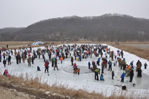 A pond that has ice on it with over hundred people fishing. The bluffs are in the background. 