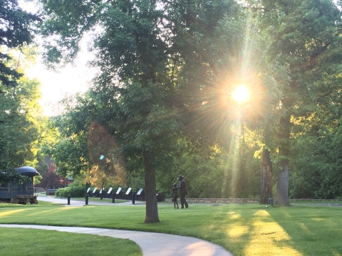 Morning sunlight streams through green trees and highlights a statue and manicured lawns.