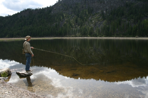 Man casting a fishing rod into a glassy lake showing the reflection of forested hills. 