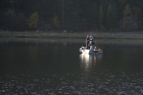 Two people stand in a boat fishing on a small lake surrounded by pine trees. 
