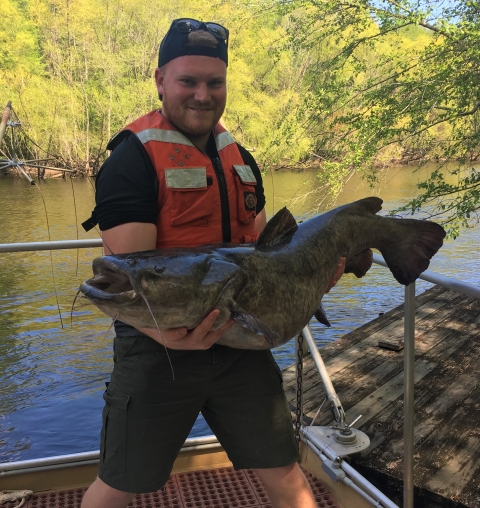 A man in a lifejacket on the bow of a boat is holding a large flathead catfish in his arms.