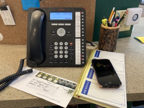 A land line phone, cell phone, letter, pencil holder with pens and pencils ,and a message pad, all on a desk. 