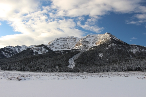 A mix of early morning sun, puffy white clouds, and blue skies over snowy mountains is shown.